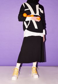 Czarny i biały sweter dziany z geometricznym wzorem, nałożony na czarną spódnicę. Białe trampki z żółtymi sznurowadłami. Modelka trzyma pomarańcze.