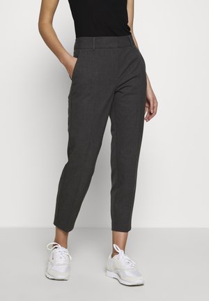 Vrouw draagt donkergrijze cropped broek, zwart mouwloos topje en witte sneakers, staand met één hand in de zak.