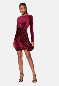 Bordó bársonyruha hosszú ujjakkal, csomózott részlettel a deréknál és légies szoknyával. A cipők fekete színűek, bokapánttal.