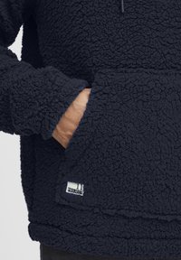 Mørkeblå fleece-pullover med teksturert overflate, med lomme foran og logo-patch. Mansjettene er brede, og kanten er lagdelt.