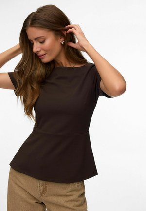 Kvinde med langt brunt hår, der retter på håret, iført mørk kortærmet top og beige bukser mod en ensfarvet hvid baggrund.