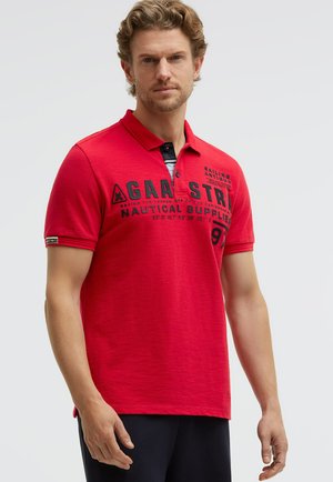 Mann mit lockigem Haar, der ein rotes Poloshirt mit schwarzem Text und Grafiken trägt, vor einem schlichten hellen Hintergrund stehend.