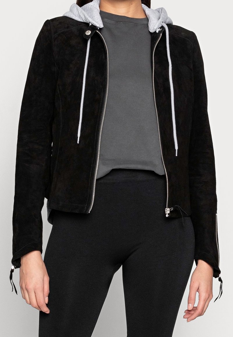 Femme portant une veste en daim noire à fermeture éclair avec un sweat à capuche gris en dessous, associée à des leggings noirs et un haut gris foncé.