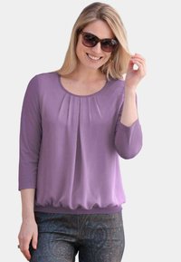 Lila langärmliges Oberteil mit Rundhalsausschnitt und gerafftem Saum, mit dezenten Falten vorne. Kombiniert mit gemusterten blauen Hosen.
