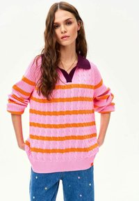 Młoda kobieta o długich brązowych włosach, ubrana w różowy sweter z pomarańczowymi paskami oraz niebieskie dżinsy z małym kwiatu haftem, stojąca z rękami w kieszeniach.