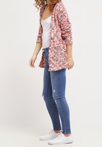 Röd, rosa och vit mönstrad cardigan med trekvartsärmar, bärs över en vit linne och blå tajta jeans, ihop med vita sneakers.