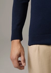 Navyblauer Strickpullover mit gerippten Bündchen, kombiniert mit hellbeigen, strukturierten Hosen. Nahaufnahme einer Hand, die an der Naht ruht.