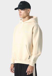 Crèmekleurige hoodie met een voorzak en een klein logo. Gladde stof, relaxte pasvorm, met ribgebreide manchetten. Draagt een zwarte pet.