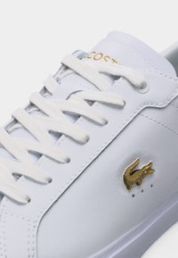 Άσπρο δερμάτινο αθλητικό παπούτσι με άσπρα κορδόνια, χρυσό λογότυπο Lacoste στο πλάι και χρυσό όνομα της μάρκας στη γλώσσα του παπουτσιού.