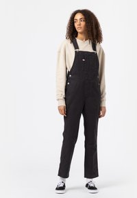 Svarta bomullsbyxor med justerbara axelband, framficka och sidoknappar, matchade med en beige sweatshirt och svarta sneakers.
