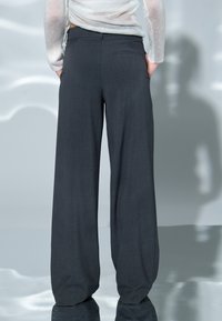 Leveälahkeiset, tummanharmaat housut, jotka on valmistettu teksturoidusta kankaasta, jossa on sileä viimeistely. Housuissa on takatasku ja hillitty räätälöinti.