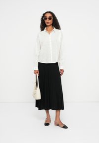 Biała bluzka z guzikiem i subtelnym wzorem, czarna, plisowana długa spódnica, beżowa torebka oraz dwukolorowe baleriny; osoba nosi okulary przeciwsłoneczne.