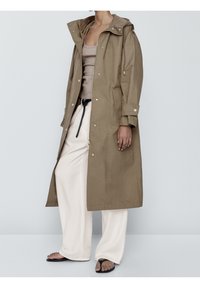 Beige long waterproof coat with a hood, featuring snap buttons, drawstring waist, and side pockets, worn over cream wide-leg pants.