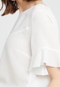 Weiße Bluse mit kurzen Ärmeln aus leichtem Stoff, runder Halsausschnitt und Rüschensäumen, mit einer glatten, leicht durchsichtigen Textur.