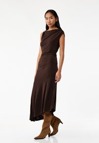 Brun, ærmeløs kjole med draperet design og samlet talje. Teksturen er glat, kombineret med knæhøje, brune ruskinds stiletto støvler.