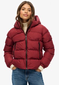 Giacca a vento rossa con cappuccio, dotata di zip frontale, polsini elasticizzati e tasche laterali. Il design trapuntato aggiunge texture e isolamento.