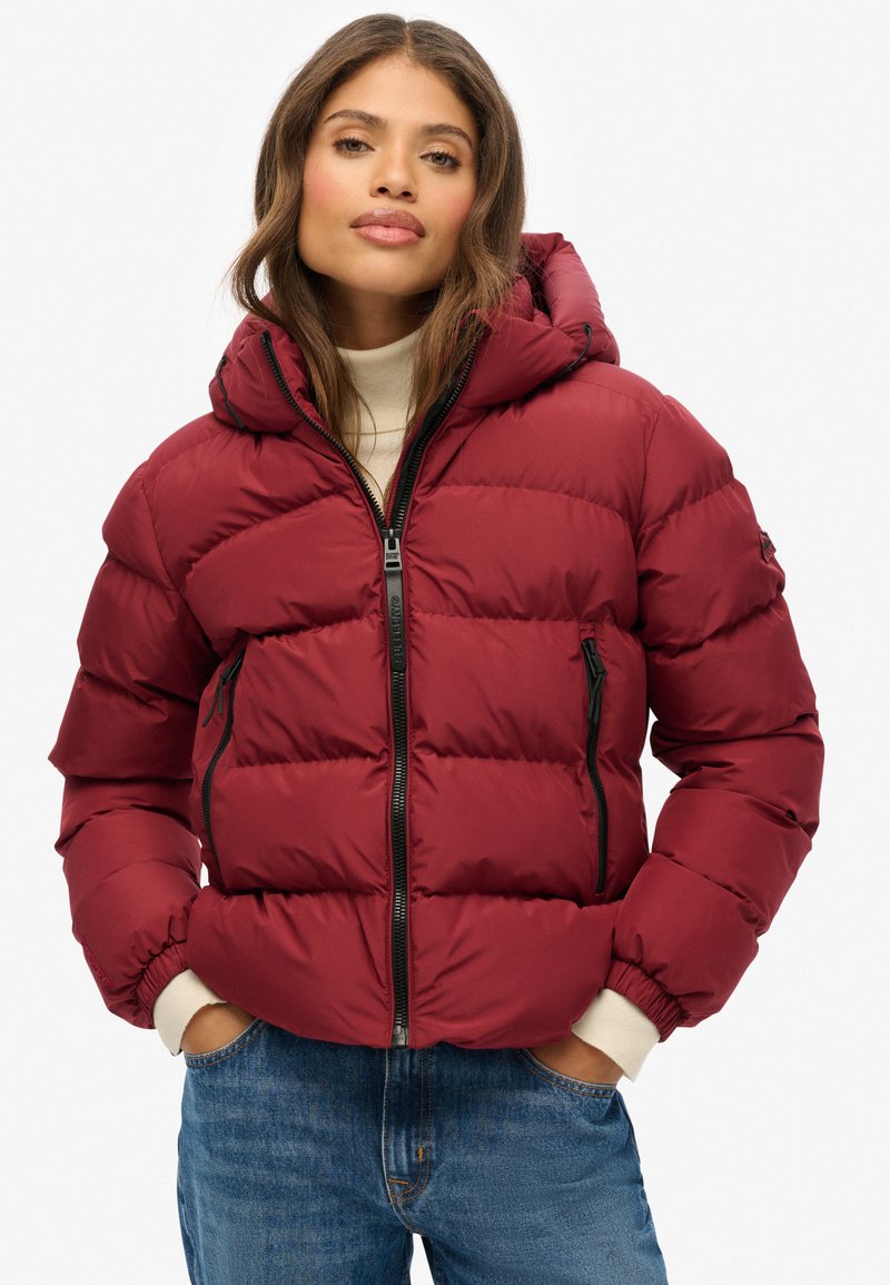 Giacca a vento rossa con cappuccio, dotata di zip frontale, polsini elasticizzati e tasche laterali. Il design trapuntato aggiunge texture e isolamento.