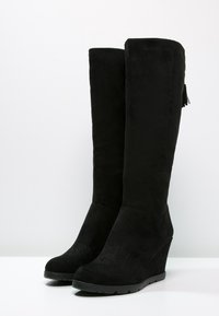 Botas de ante negras hasta la rodilla con tacón de cuña, que cuentan con un sutil patrón en relieve cerca del dedo del pie y un adorno de borlas en la parte superior.