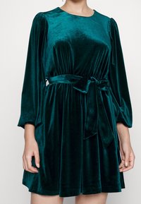 Siniharmaa samettinen mekko, jossa on pitkät hihat, pyöreä kaula-aukko ja solmittava vyötärönauha. Sileä pinta ja hieman levenevä helmankuvio.