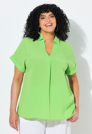 Femme aux cheveux noirs bouclés souriante, portant un chemisier vert vif à manches courtes avec col et un pantalon blanc, debout devant un fond clair.