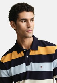 Koszulka polo z poziomymi paskami w kolorach granatowym, żółtym i jasnoniebieskim. Posiada kołnierzyk oraz guziki z przodu, a także akcent logo.