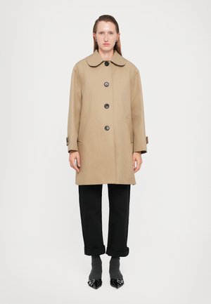 Cappotto beige in lana con colletto rotondo, dotato di grandi bottoni neri e tasche laterali. Indossato sopra pantaloni neri e scarpe nere a punta.