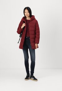 Doudoune bordeaux avec capuche, design matelassé texturé, associée à un jean foncé et des baskets noires avec des rayures blanches et des semelles en caoutchouc.