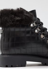 Bota de cuero negro con textura acolchada, cuello de piel sintética, ojales plateados y cordones. Suela de goma con patrón de dibujo para tracción.