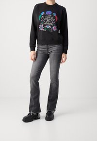 Sudadera negra con un diseño gráfico colorido que presenta pájaros y patrones. Combinada con jeans acampanados grises y zapatos negros de suela gruesa.