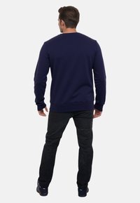 Felpa in cotone blu navy con maniche lunghe, colletto tondo e polsini a coste. Indossata con pantaloni neri e calzature nere, vista da dietro.