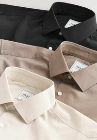 Trois chemises à col en noir, taupe et crème, affichant un tissu lisse, des fermetures à boutons et des étiquettes de marque portant "NEXT Tailored."
