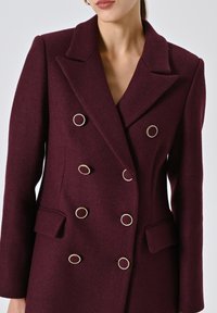 Cappotto bordeaux doppiopetto in tessuto strutturato. Presenta bottoni circolari dorati, colletto a punte e due tasche anteriori.