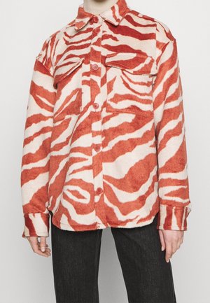 Langärmlige Jacke mit Knopfleiste, rot-weißen Zebramustern und zwei aufgesetzten Taschen vorne, getragen über einer schwarzen Hose.