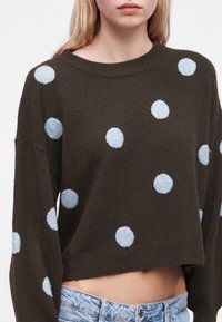 Jeune femme portant un pull court marron foncé avec de gros pois bleu clair et un jean bleu clair, debout devant un fond uni.
