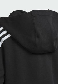 Sort hættetrøje med struktureret stof, der har en stor hætte og tre hvide striber langs den venstre skuldersøm. Minimalistisk design.