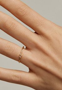 Anillo de oro con una banda delgada y tres pequeñas piedras preciosas claras engastadas en línea. La mano presenta una textura de piel natural y un tono bronceado claro.