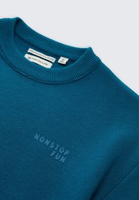 Türkisfarbenem Pullover aus umweltfreundlicher Viskose, mit geripptem Rundhalsausschnitt und geprägtem "NONSTOP FUN"-Text auf der Vorderseite. Glatte Textur.