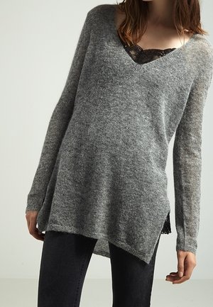 Femme portant un pull gris ample à col en V sur un top en dentelle noire et un pantalon noir, debout devant un fond clair uni.