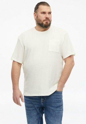 Man with beard wearing a cream short-sleeve shirt with chest pocket and blue jeans, standing with one hand in pocket, looking right.