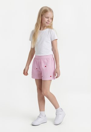 Jeune fille portant un t-shirt blanc, un short rose avec de petits cœurs rouges, des chaussettes blanches et des baskets blanches, debout sur un fond blanc.