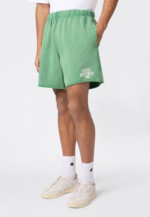 Mężczyzna w zielonych szortach z napisem „Pacific Northwest”, białych skarpetkach Champion i kremowych tenisówkach, stoi z jedną ręką w kieszeni.