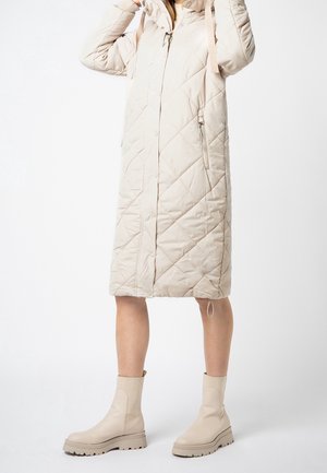 Femme portant un manteau long matelassé beige avec capuche et des bottines beiges, ajustant la capuche avec les deux mains.
