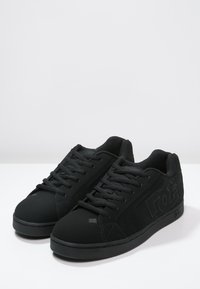 Sorte sneakers med en glat tekstiloverdel, rund tå, flade sorte snørebånd og logo-detaljer på siden. Gummisål med subtilt mønster.