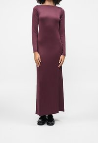 Femme portant une robe longue, ajustée, à manches longues, violet foncé, avec un décolleté bateau et des chaussures noires brillantes, debout devant un fond blanc.