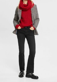 Karierter Blazer, dicker roter Strickpullover, schwarze Jeans und glänzende schwarze Schuhe. Der Blazer hat ein tailliertes Design; der Pullover hat einen Schalkragen.