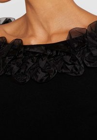 Svart topp med en volangkrage i genomskinlig svart tyg, med en slät textur och en åtsittande silhuett.