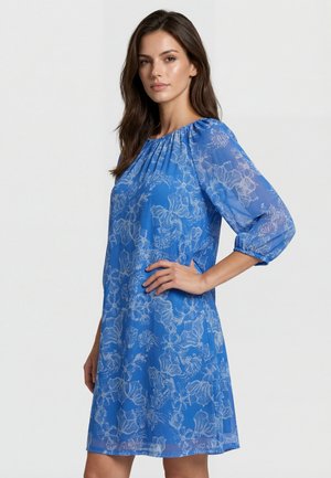 Kvinde med langt brunt hår iført en blå knælængde kjole med hvide blomsterprint og trekvart gennemskinnelige ærmer, poserer med den ene hånd på hoften.