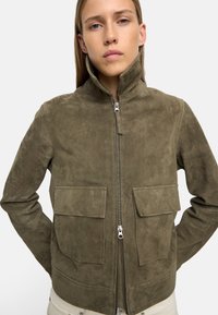 Zielono-oliwkowa skórzana kurtka z wysokim kołnierzem, zamkiem błyskawicznym z przodu i dwiema dużymi kieszeniami na piersi. Gładka tekstura i czyste linie.