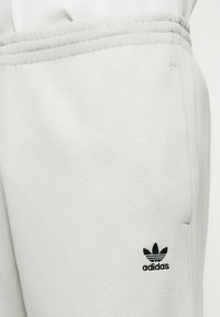 Gaiši pelēkas sporta bikses ar elastīgu jostu un sānu kabatu, ar mazu melnu Adidas logotipu blakus kabatai.