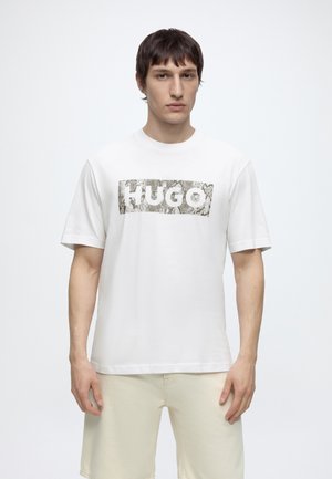Nuori mies, jolla on yllään valkoinen lyhythihainen paita, jossa on käärmekuvioinen logo "HUGO", ja vaaleanbeiget shortsit, seisoo yksiväristä taustaa vasten.